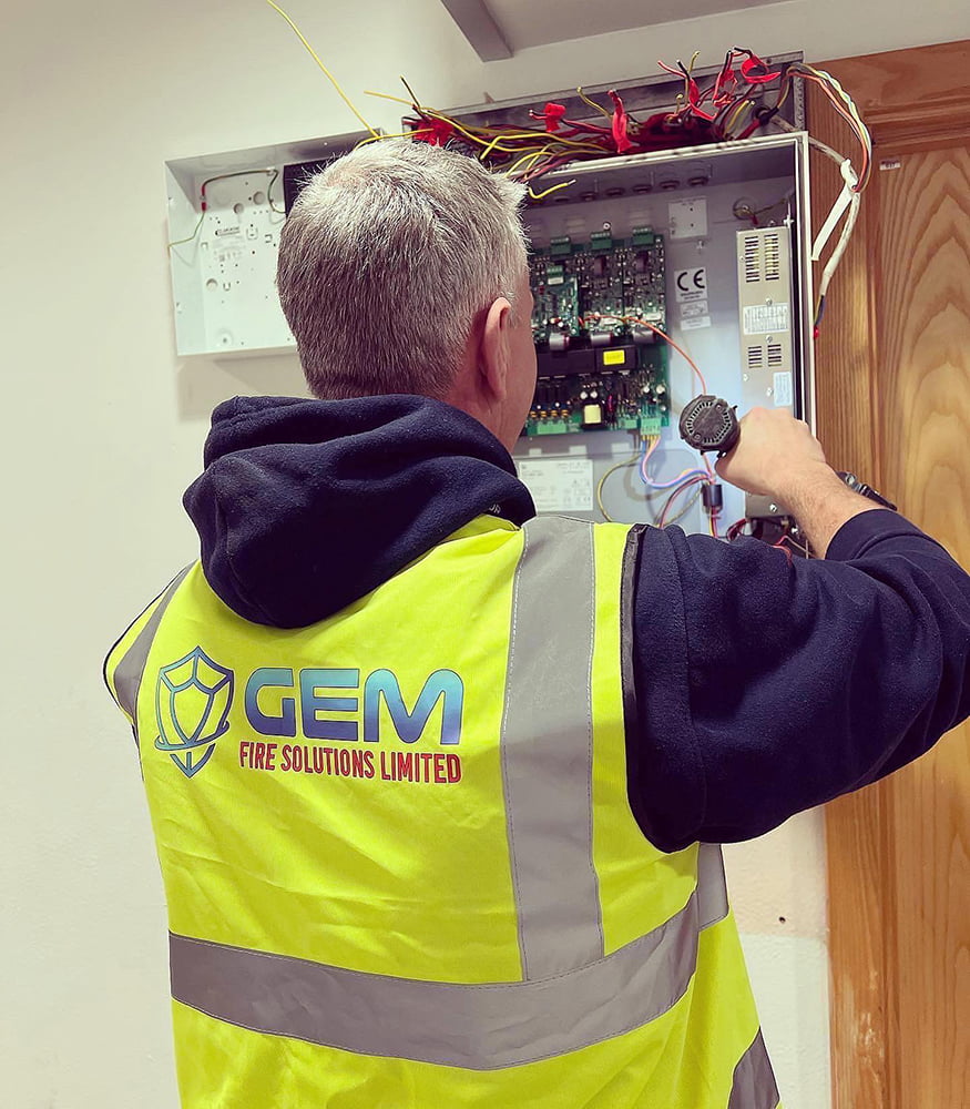 GEM Fire Solutions' employee installing fire alarm. Wearing yellow vest with reflectors and company's name